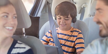 A man and woman glance knowingly at each other as their son contentedly listens to headphones in the sunny backseat, protected from harmful UV rays.
