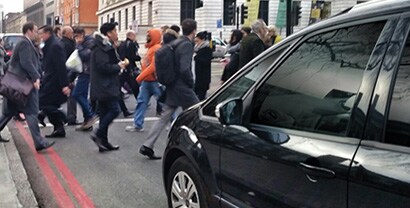 Crowds of people walk past a car with black, reflective window films.
