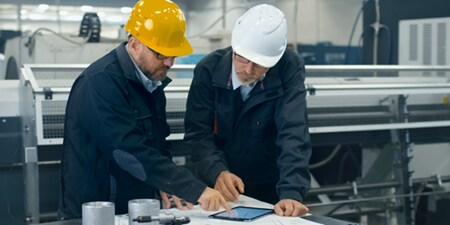 Two people wearing hard hats look at a tablet resting on a table.
