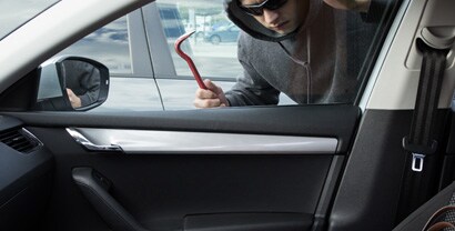 A man wearing a black hoodie and carrying a crowbar pauses as he leans down in front of a car window to try to see what’s inside.
