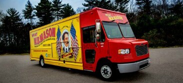 A van with a brightly colored vehicle wrap displays a business name and an image of a man with two American flags.
