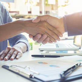 Two people shake hands over a table with pens, a clipboard and a laptop