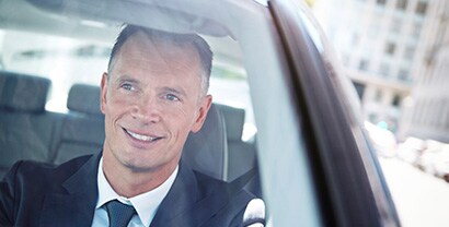 A professional man smiles as he comfortably drives without sunglasses on a sunny day.

