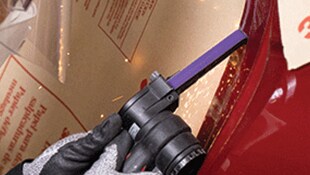 A gloved hand using a handheld power tool with a narrow abrasive belt on a metal surface
