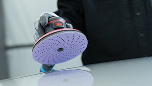 A gloved hand holding a 3M orbital sander with a perforated sanding disc
